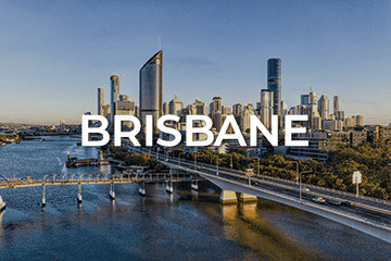 Aerial view of Brisbane with the city's skyline in the background. The river flows beneath a prominent bridge, reflecting the vibrant energy of a city bustling with opportunities, like a business for sale. Clear blue sky suggests a sunny day. 'BRISBANE' is overlaid in bold white letters.