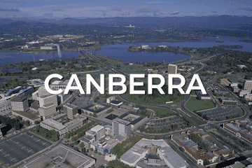 Aerial view of Canberra, showcasing prominent buildings, roads, and green spaces. Large text 'CANBERRA' overlays the center of the image, perhaps highlighting a thriving city with attractive business for sale opportunities. A river and distant hills grace the background.